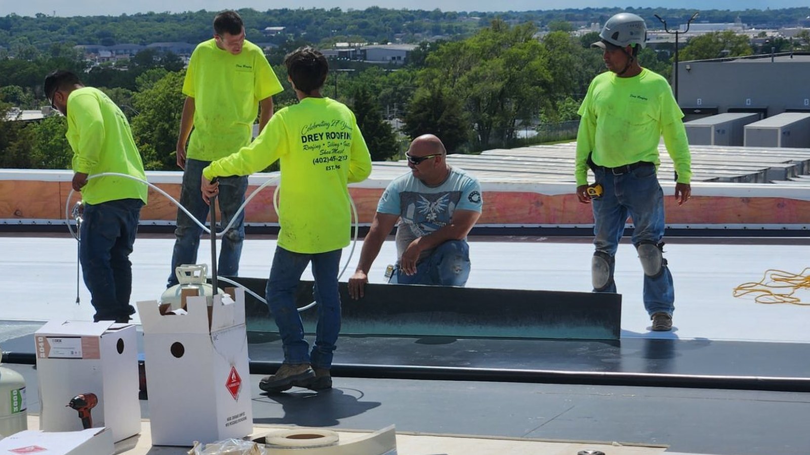 Roofing crew installing single-ply membrane on a commercial flat roof with QDEK adhesive canisters staged nearby.